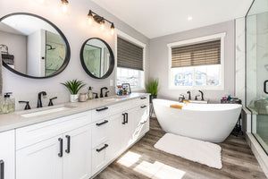 Light grey walls, white double vanity with light grey countertop. White freestanding tub and black plumbing fixtures. Black round mirrors. Brown vinyl floor.